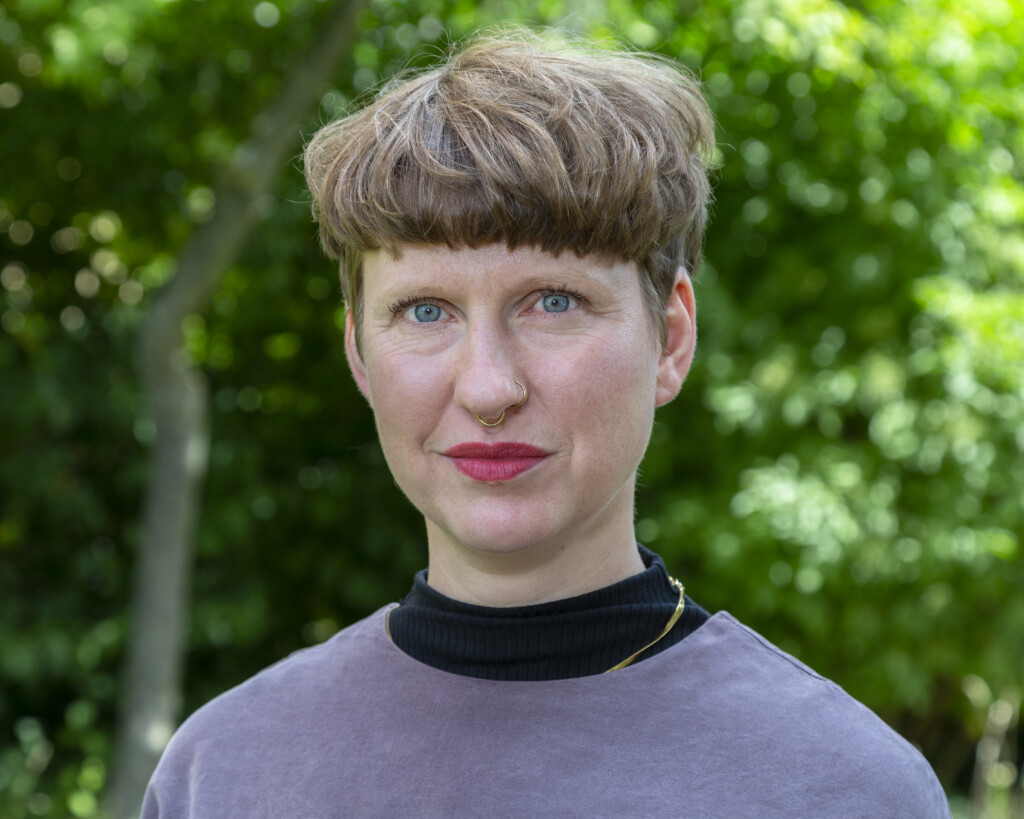 Porträtfoto einer Person mit kurzem, welligem, hellbraunem Haar. Sie trägt ein graublaues Oberteil mit hohem schwarzen Kragen und hat einen Nasenring mittig uns einen links. Ihr Blick ist freundlich, und sie hat ein dezentes Lächeln. Der Hintergrund ist unscharf und zeigt grüne Blätter.