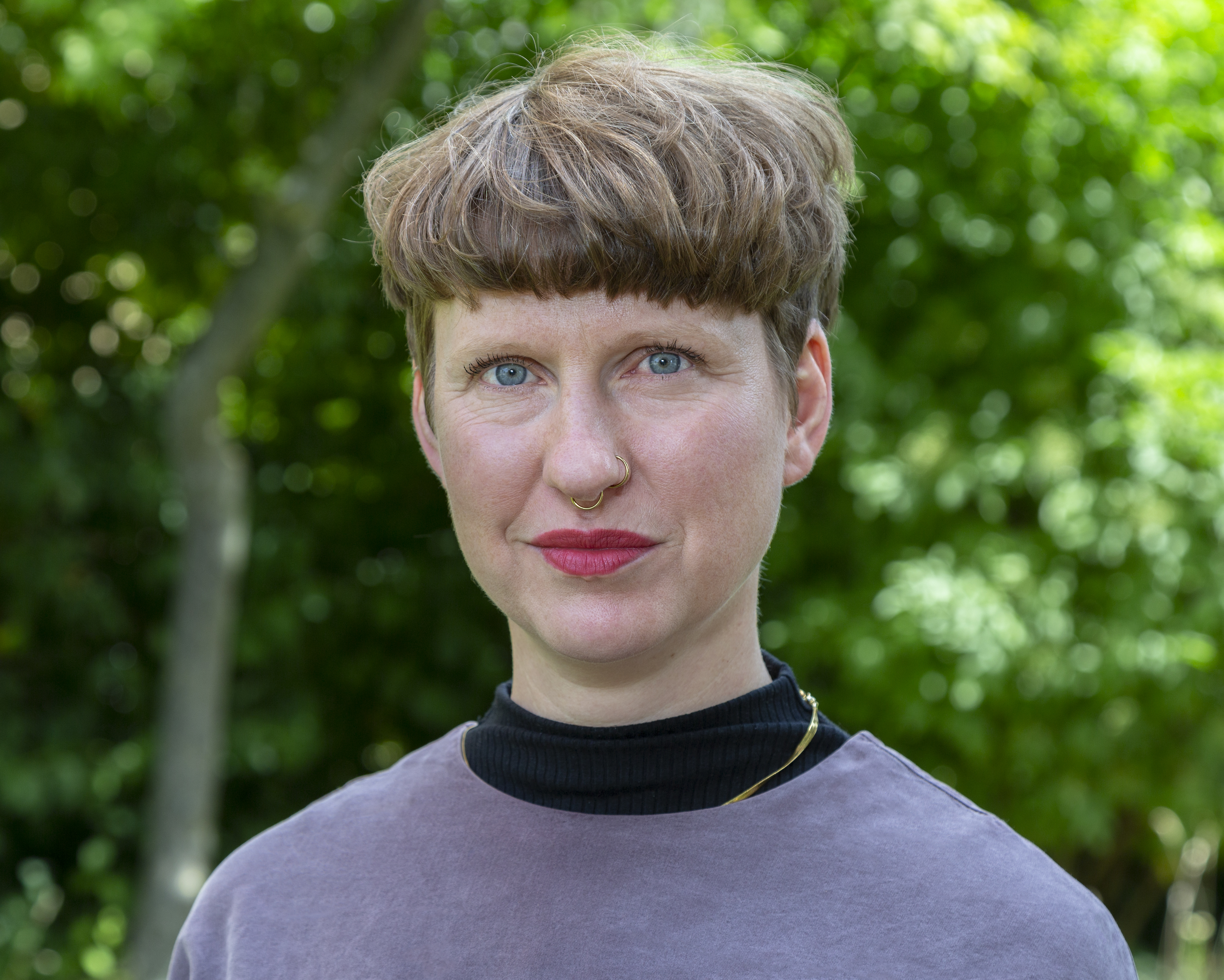 Porträtfoto einer Person mit kurzem, welligem, hellbraunem Haar. Sie trägt ein graublaues Oberteil mit hohem schwarzen Kragen und hat einen Nasenring mittig uns einen links. Ihr Blick ist freundlich, und sie hat ein dezentes Lächeln. Der Hintergrund ist unscharf und zeigt grüne Blätter.