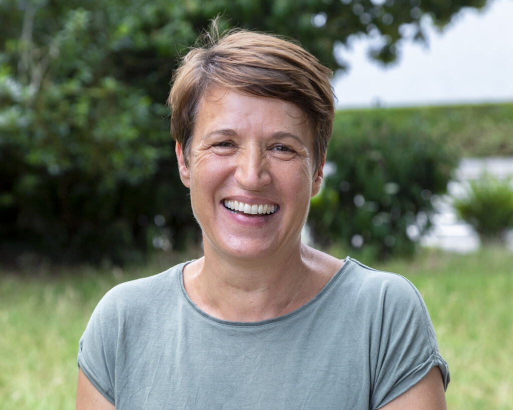 Porträt einer Frau mit sehr dunkelbraunen kurzen Haaren, die locker in die Stirn fallen. Sie trägt ein blau-grünes Oberteil. Sie lacht freundlich in die Kamera, während im Hintergrund grüne Blätter zu sehen sind.
