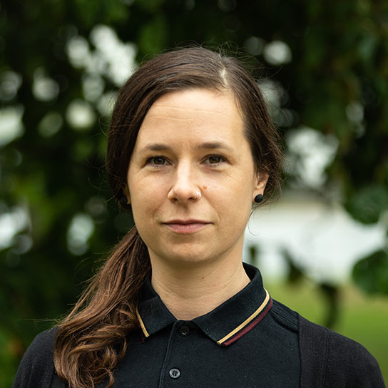 Porträt einer Frau mit dunkelbraunen Haaren, die nach hinten gebunden sind. Der Pferdeschwanz liegt auf der rechten Schulter. Sie trägt ein Nasenpiercing, schwarze Ohrringe und ein schwarzes Poloshirt und eine schwarze Strickjacke. Sie lächelt freundlich in die Kamera, während im Hintergrund grüne Blätter zu sehen sind.