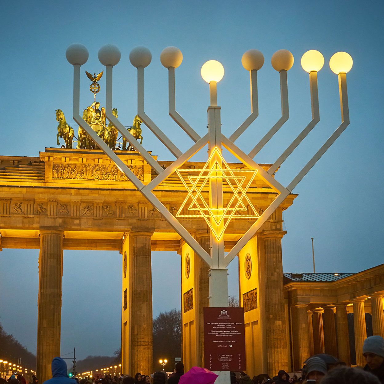 Eine große Chanukkia (jüdischer Leuchter mit acht Armen) mit leuchtendem Davidstern steht vor dem Brandenburger Tor in Berlin. Die Lichter der Chanukkia sind teilweise entzündet. Im Hintergrund erstrahlt das Brandenburger Tor mit seiner berühmten Quadriga, während sich zahlreiche Menschen in winterlicher Kleidung davor versammeln. Der Himmel ist dunkelblau.