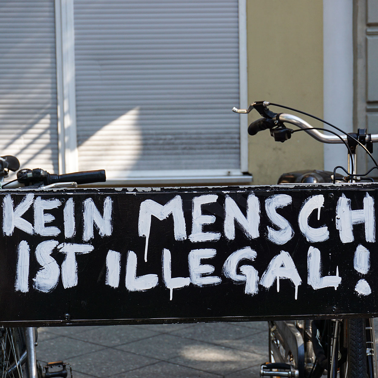 Kein Mensch ist illegal steht in großen weißen gemalten Buchstaben auf der schwatzen Rückseote einen Fahrradständers. Im Hintergrund ist eine Häuserfassade mit runtergelassenen Rolläden.