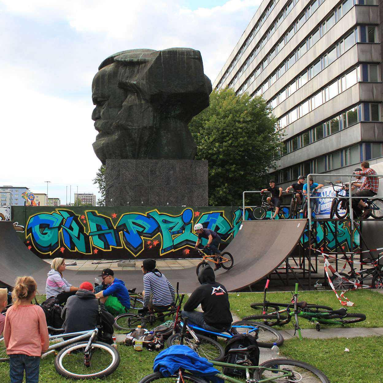 Eine Gruppe Jugendlicher auf BMX-Rädern und Skateboards nutzt eine Halfpipe mit Graffiti vor der monumentalen Karl-Marx-Büste in Chemnitz, auch als „Nischel“ bekannt. Die Szene kombiniert urbanen Sport mit historischer Kulisse, während im Hintergrund moderne Gebäude sichtbar sind.