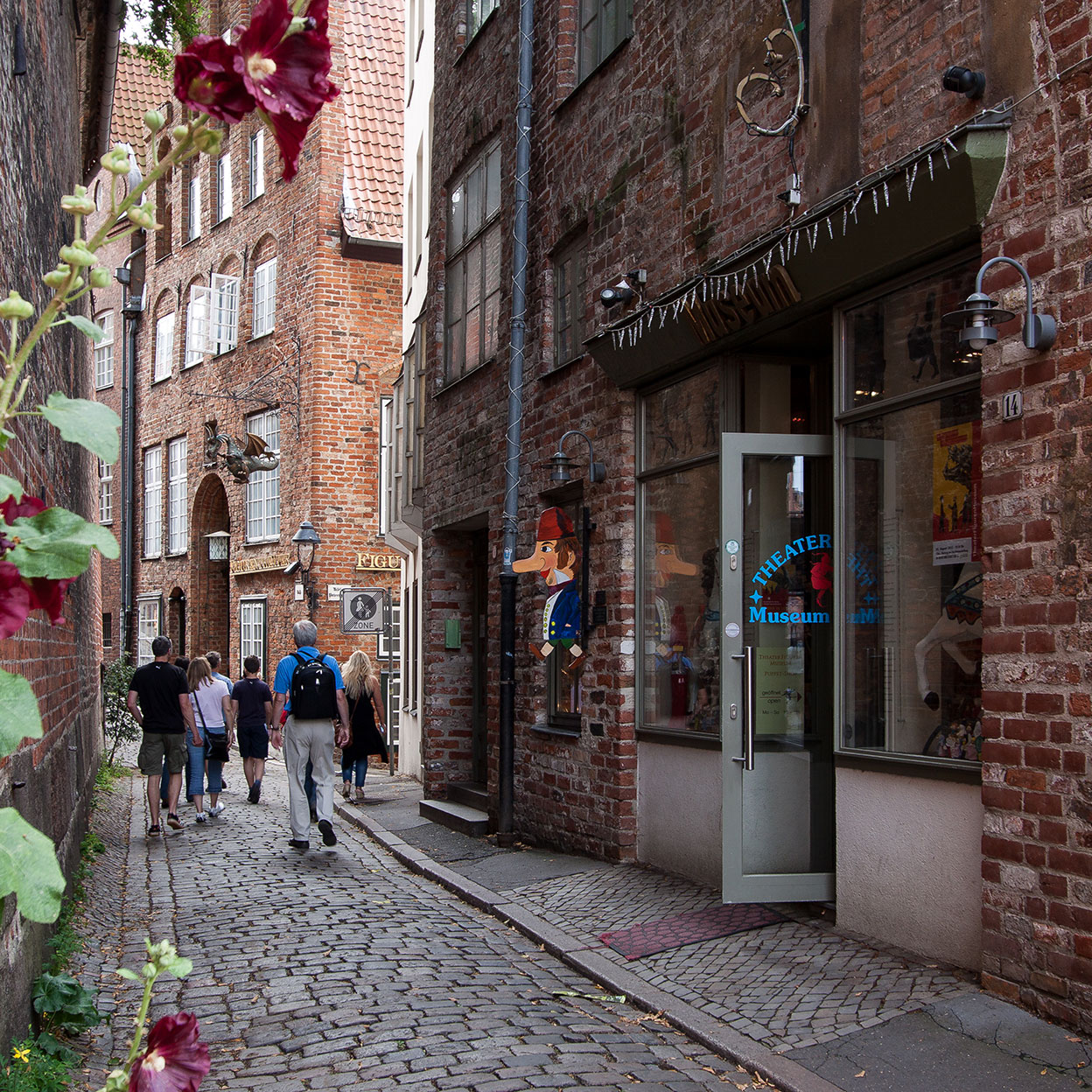 Eine malerische Gasse in Lübeck, gesäumt von roten Backsteinhäusern. Im Vordergrund ranken Stockrosen am Rand der Kopfsteinpflasterstraße. Besucher schlendern entlang der Straße, vorbei an einem kleinen Museum mit Schaufenstern und offenen Türen, das lebhafte Puppentheater-Requisiten zeigt. Die historische Atmosphäre spiegelt die hanseatische Architektur wider und lädt zum Erkunden ein.