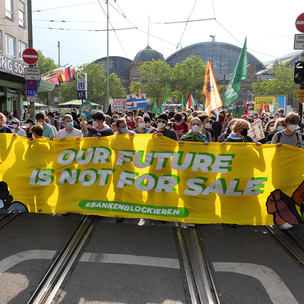 Eine Demonstration in Frankfurt. Im Hintergrund ist der Hauptbahnhof. Mit einer großen gelben Banneraufschrift 'Our Future is Not for Sale' und dem Hashtag #BankenBlockieren. Viele Menschen tragen Masken und halten Plakate hoch. Im Hintergrund sind Bäume und ein historisches Bahnhofsgebäude zu sehen.