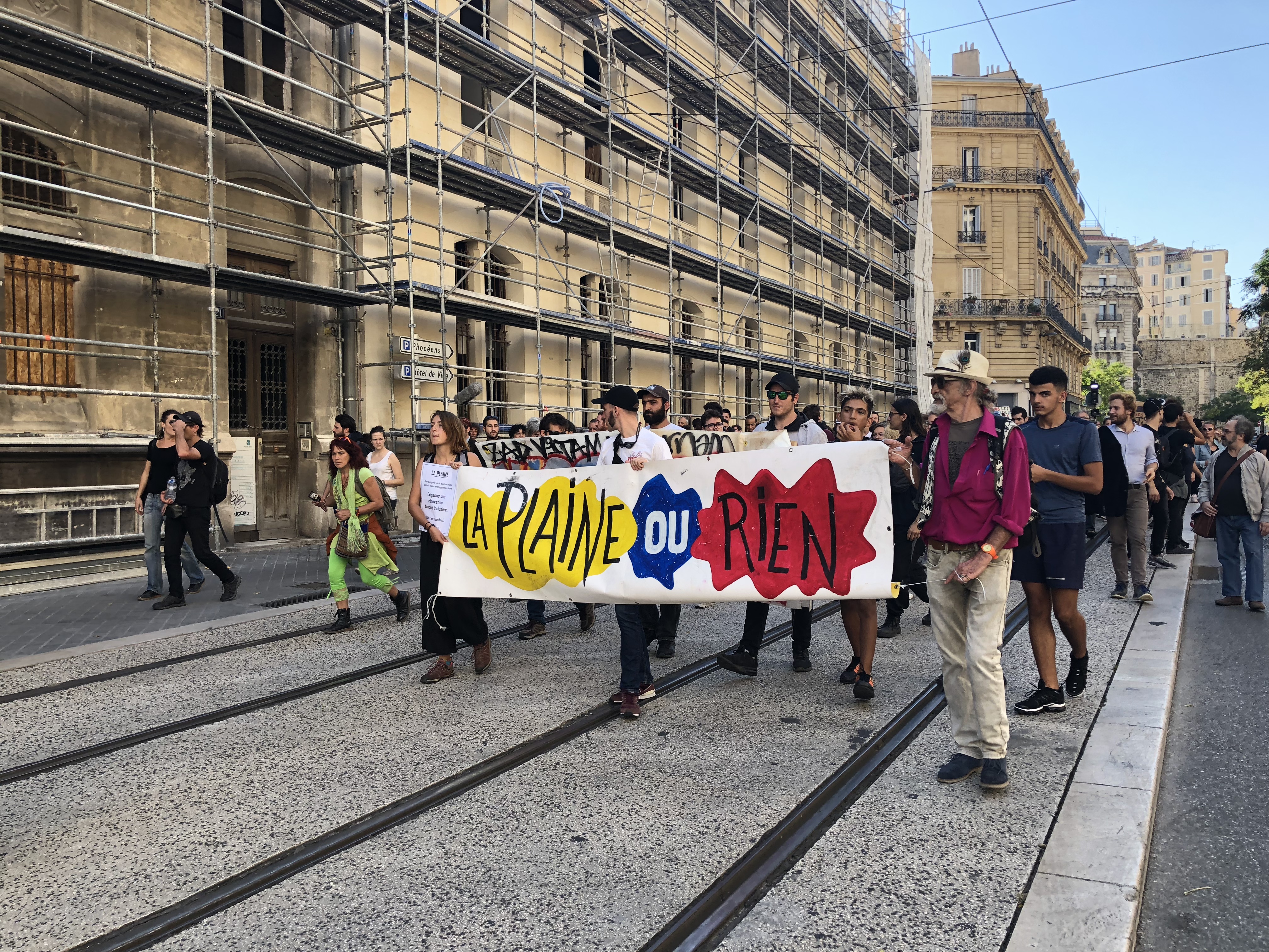 Menschen demonstrieren auf einer Straße in Marseille. Sie halten ein Banner mit der Aufschrift ‚La Plaine ou rien‘ in leuchtenden Farben. Umgeben von alten Gebäuden und Gerüsten, spiegelt die Szene den Ausdruck von Protest und Gemeinschaft wider.