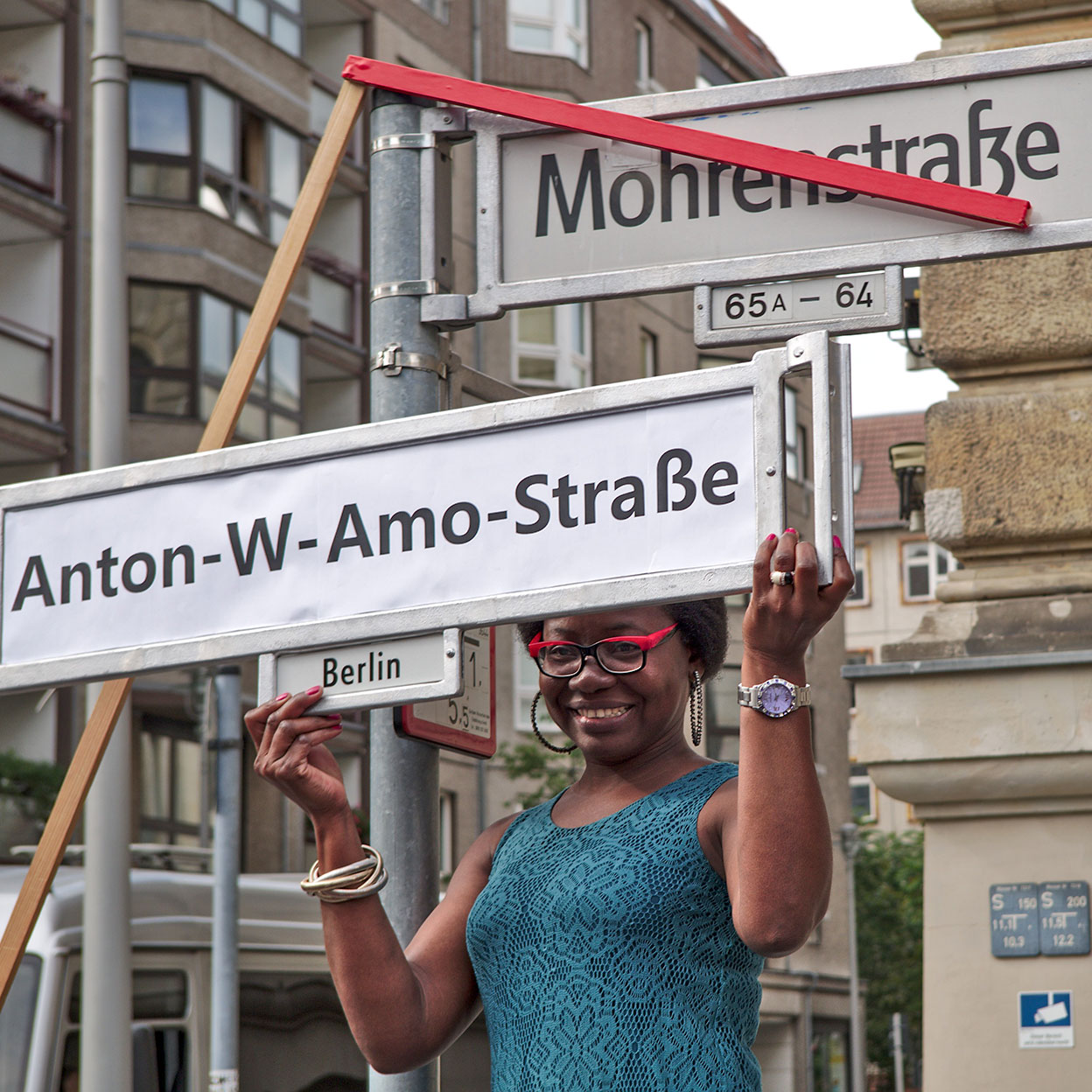 Eine Schwarze Frau mit roter Brille und einem türkisfarbenen Kleid steht lächelnd vor einem Straßenschild. Sie hält ein neues Schild mit der Aufschrift 'Anton-W.-Amo-Straße' über das alte Straßenschild 'Mohrenstraße', das mit rotem Klebeband durchgestrichen ist. Im Hintergrund sind Berliner Straßenschilder und Wohngebäude zu sehen. Die Szene symbolisiert eine kritische Auseinandersetzung mit kolonialen Straßennamen und eine Initiative zur Umbenennung.