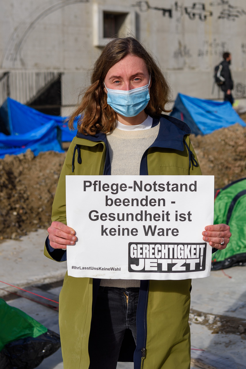 Eine Person mit blauem Mundschutz guckt ernst in die Kamera und hält ein Schild hoch auf dem steht: Pflege-Notstand beenden - Gesundheit ist keine Ware. Gerchtigkeit jetzt! #Ihr lasst uns keine Wahl. Im Hintergund sind kleine Zelte aus blauer Plane