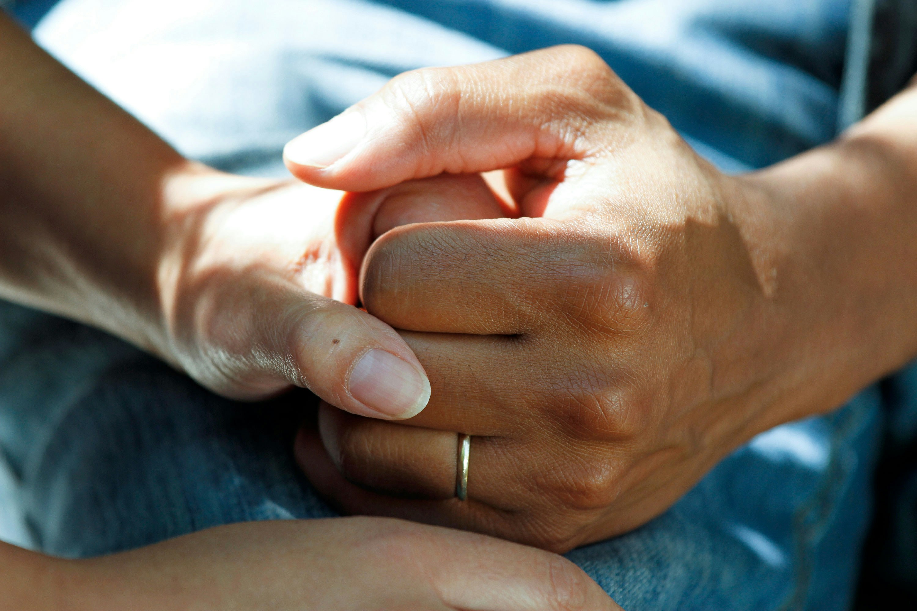 Auf einer blauen Jeans greifen die Hände von zwei Personen ineinander. An der Hand der einen Person ist ein Ring.