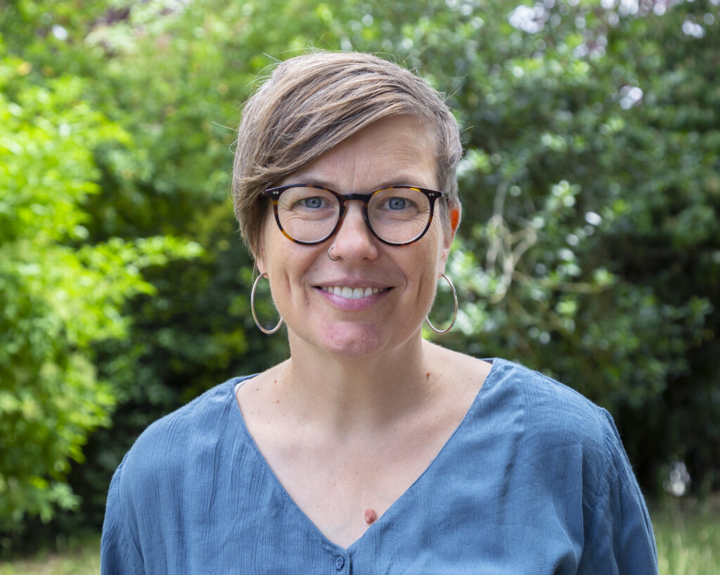 Porträt einer Frau mit kurzen braunblonden Haaren, die nach rechts fallen. Sie trägt eine Brille mit dunklem Rand, Kreolen Ohrringe und sie hat ein kleines Nasenpiercing rechts. Sie hat ein blaues Oberteil an. Sie lächelt freundlich in die Kamera, während im Hintergrund grüne Blätter zu sehen sind.