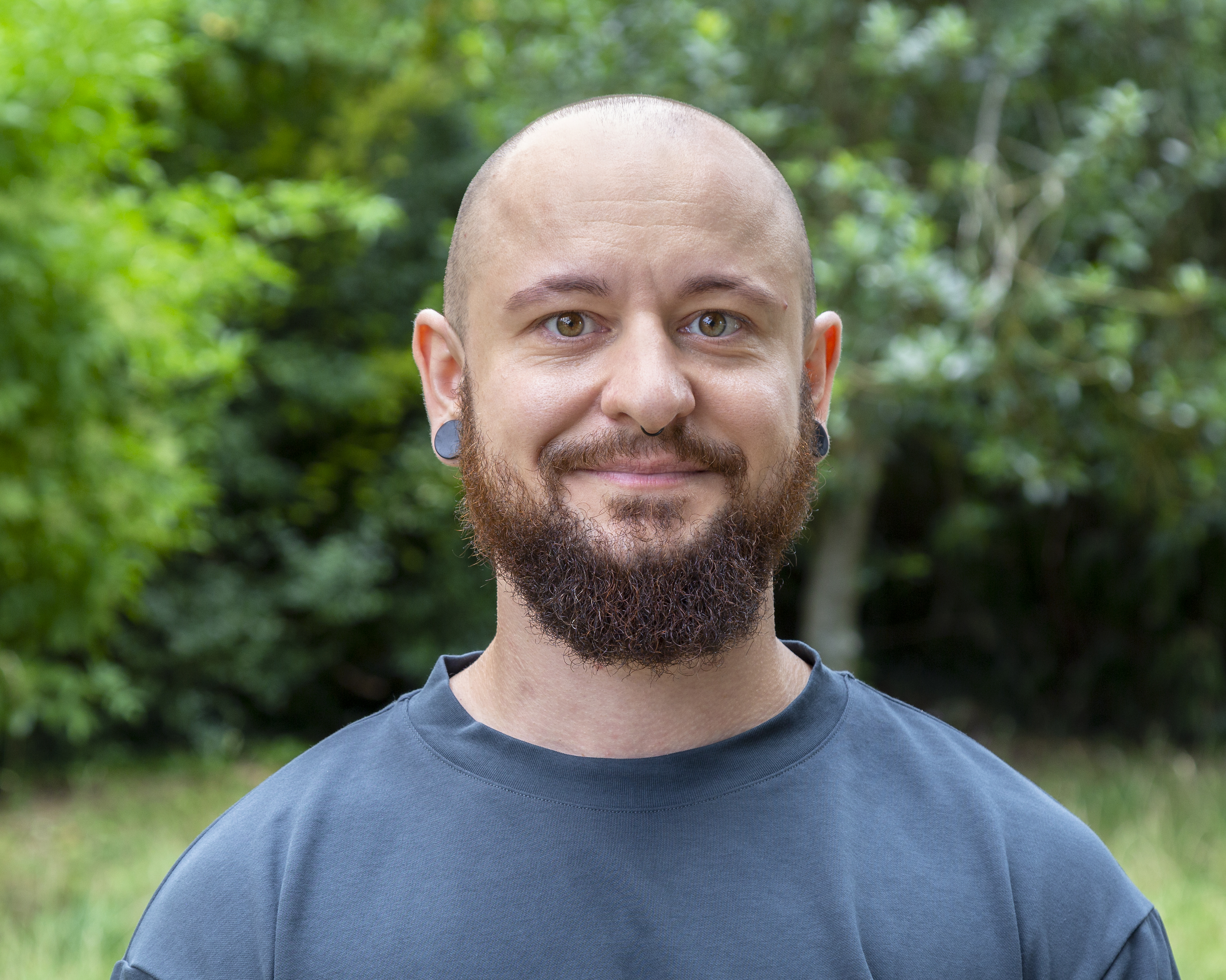 Porträt einer lächelnden Person mit rasiertem Kopf und dunklem Vollbart. Sie trägt ein petrolfarbenes T-Shirt und hat Tunnel-Piercings in den Ohren. Im Hintergrund sind grüne Blätter zu sehen.