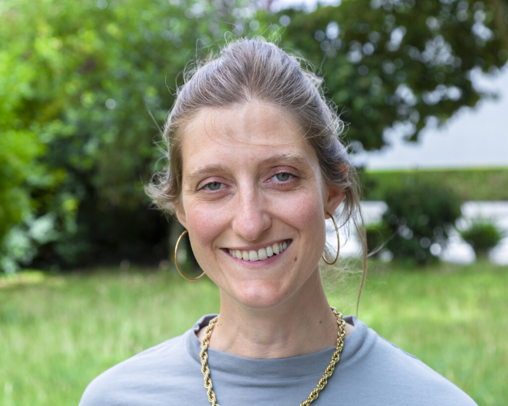 Porträt einer Frau mit braunen Haaren, die nach hinten gebunden sind. Sie trägt eine goldene Kette, goldene Ohrringe und ein blau graues Oberteil. Sie lächelt freundlich in die Kamera, während im Hintergrund grüne Blätter zu sehen sind.