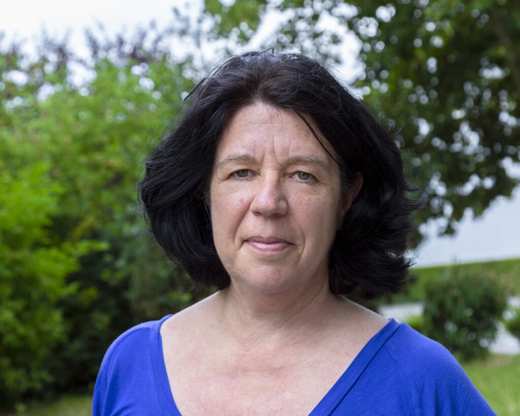 Porträt einer Frau mit kinnlangen schwarzen Haaren. Sie trägt eine ein leuchtend blaues Oberteil. Sie lächelt freundlich in die Kamera, während im Hintergrund grüne Blätter zu sehen sind.