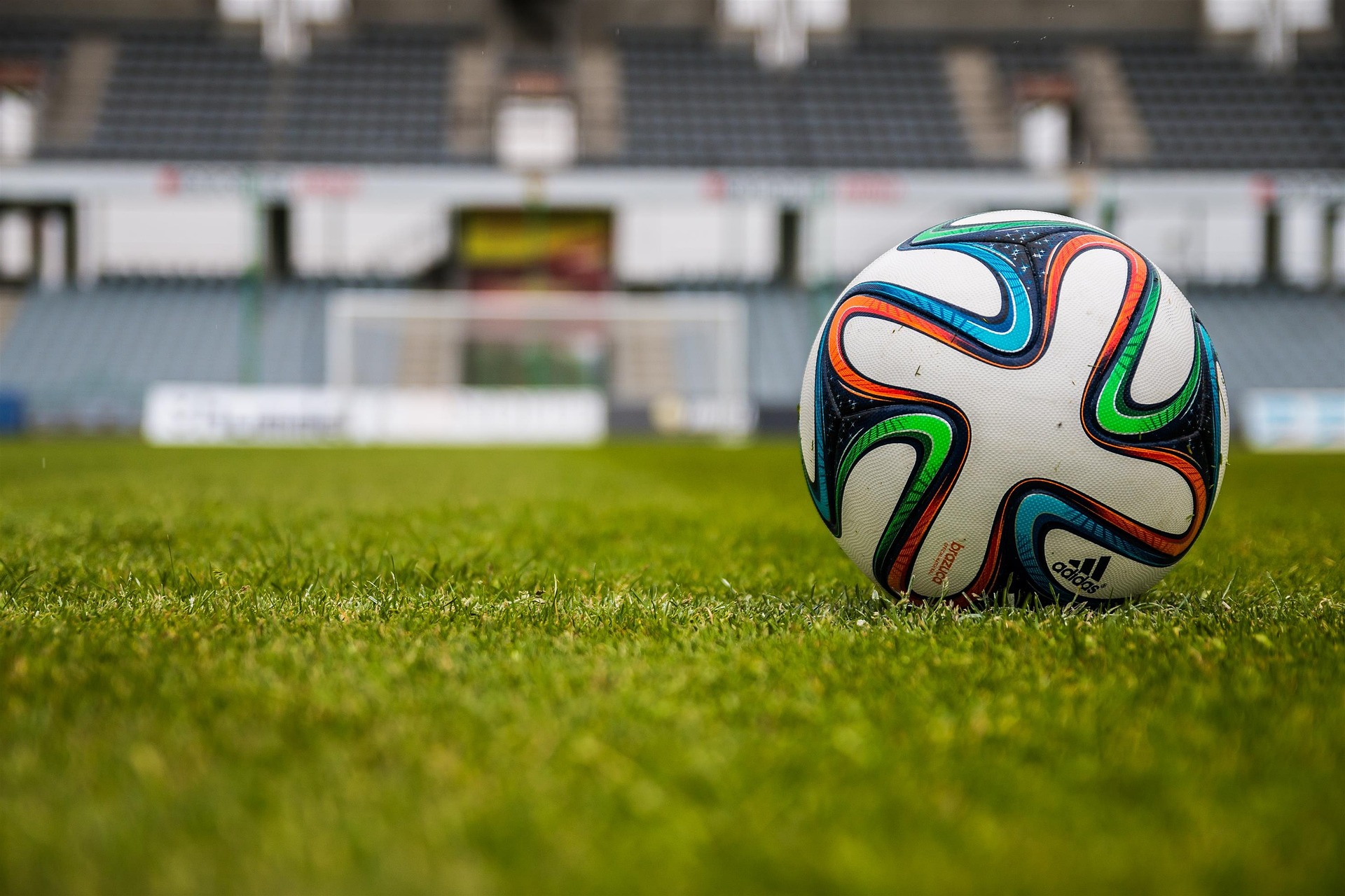 Ein Fußball liegt auf Rasem. Im Hintergrund sind die Tribünen eines Stadions.