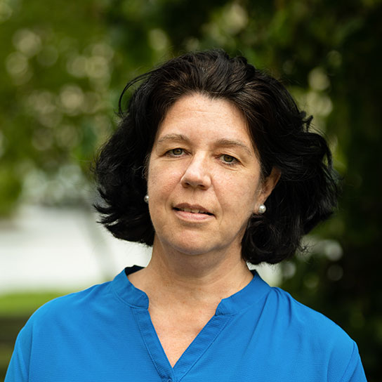 Porträt einer Frau mit kinnlangen schwarzen Haaren.. Sie trägt eine Perlenohrringe und ein leuchtend blaues Oberteil. Sie lächelt freundlich in die Kamera, während im Hintergrund grüne Blätter zu sehen sind.