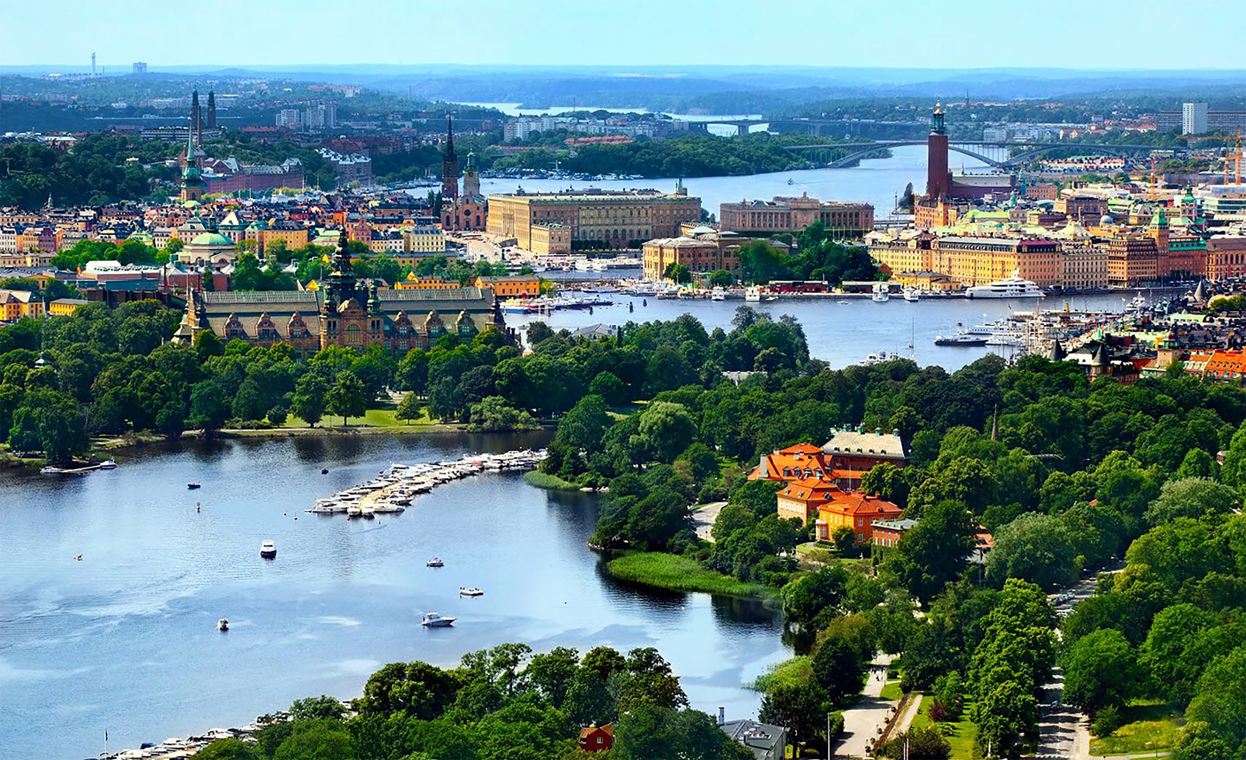 Ein Panoramablick auf Stockholm an einem sonnigen Tag, mit grünen Parks, Wasserwegen und zahlreichen Booten. Im Vordergrund liegt eine üppig bewachsene Insel, während im Hintergrund historische Gebäude, Kirchen und das Rathaus von Stockholm zu sehen sind. Die Stadt erstreckt sich entlang eines Netzwerks von Flüssen und Brücken.