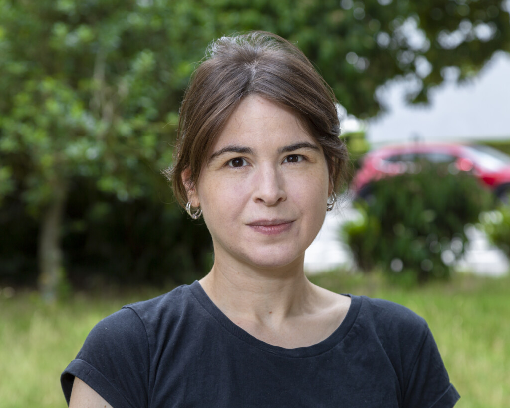 Porträt einer Frau mit braunen Haaren, die nach hinten gebunden sind. Sie trägt silberne Ohrringe und ein schwarzes Oberteil. Sie lächelt freundlich in die Kamera, während im Hintergrund grüne Blätter zu sehen sind.