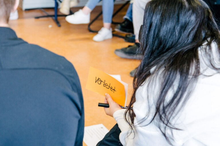 Eine Person hält eine orangefarbene Karte mit der Aufschrift 'verletzt' in der Hand und sitzt in einem Stuhlkreis mit anderen Teilnehmenden. Im Hintergrund sind Beine und Schuhe der anderen Personen zu sehen