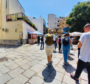 Eine Gruppe von Menschen spaziert an einem sonnigen Tag durch die Altstadt von Palermo. Sie tragen Rucksäcke und Hüte, was auf eine geführte Tour hindeutet. Links sind kleine Marktstände mit bunten Sonnenschirmen zu sehen, während im Hintergrund eine Hauswand mit Graffiti und grüne Bäume das Stadtbild prägen. Die Pflastersteine und die mediterrane Architektur verleihen der Szene eine lebendige und einladende Atmosphäre.