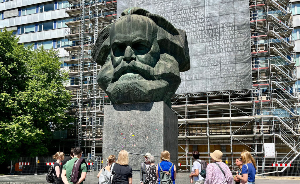 Eine Gruppe steht vor dem Karl-Marx-Kopf in Chemnitz. Vor der Gruppe steht ein Mann und erzält etwas.