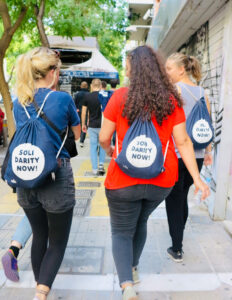 Drei junge Personen gehen eine Stadtstraße entlang und tragen dunkelblaue Turnbeutel mit der Aufschrift 'SOLIDARITY NOW!'. Vor ihnen läuft eine Gruppe weiterer Menschen. Die Umgebung ist urban, mit Graffiti an den Wänden und grünen Bäumen am Straßenrand.