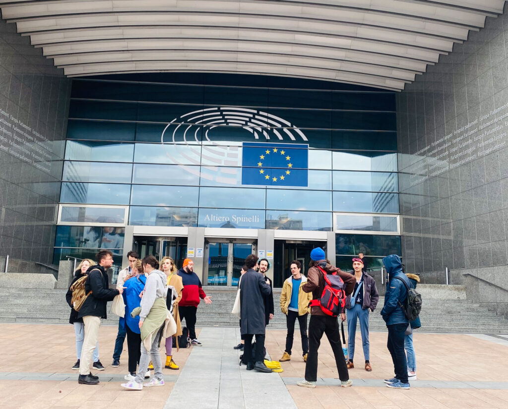 Eine Gruppe junger Menschen steht vor dem Altiero-Spinelli-Gebäude des Europäischen Parlaments in Brüssel. Über dem Eingang prangt eine große EU-Flagge, und die Glasfassade spiegelt den Himmel. Die Gruppe scheint im Gespräch zu sein, einige tragen Rucksäcke und wetterfeste Kleidung