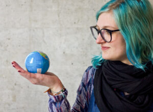 Eine Person mit türkisen Haaren und einer Brille mit schwarzen Rahmen hält auf ihrer Handfläche einen kleinen Globus. Sie guckt den Globus an und lächelt.