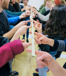 Eine Gruppe von Menschen steht sich in zwei Reihen gegenüber und hält gemeinsam mit den ausgestreckten Zeigefingern einen langen, zusammengeklappten Zollstock. Die Übung scheint Teamarbeit oder Geschicklichkeit zu fördern.