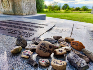 Ein Gedenkort mit einer Gedenktafel, auf der mehrere Zeilen in verschiedenen Sprachen eingraviert sind. Davor liegen bemalte und beschriftete Steine mit Botschaften wie 'Never Again' und anderen in verschiedenen Sprachen, darunter Hebräisch, Arabisch und Deutsch. Die Steine symbolisieren das Gedenken an die Opfer. Im Hintergrund erstreckt sich eine grüne Landschaft mit Bäumen und Bergen.