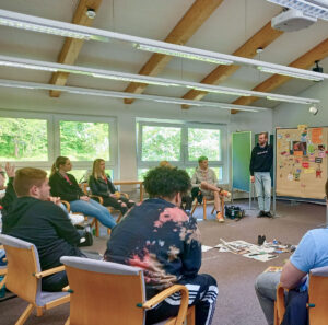 Ein Workshop in einem modernen Seminarraum mit großen Fenstern, die ins Grüne blicken. Junge Erwachsene sitzen in einem Stuhlkreis und diskutieren engagiert. Eine Person steht neben einer Pinnwand mit bunten Zetteln und Bildern, während eine Person aus der Gruppe eine Frage stellt. Auf dem Boden liegen Zeitungen und Materialien für kreative Arbeit