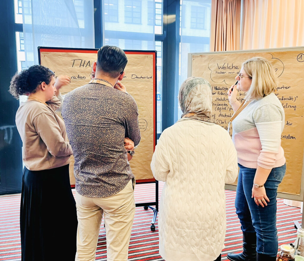 Vier Personen stehen vor zwei Pinnwänden. Die Pinnwände sind mit Papier bespannt und handbeschrieben. Die Person rechts im Bild hält einen Marker in die Luft.