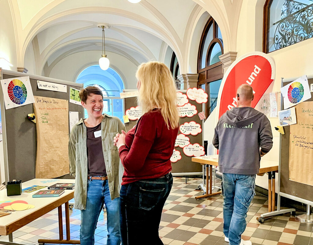 Drei Personen in einem historischen Gebäude mit hohen Bögen und großen Fenstern nehmen an einer Veranstaltung teil. Im Vordergrund unterhalten sich zwei Frauen angeregt und lachen. Im Hintergrund betrachtet ein Mann mit einer Kapuzenjacke eine Pinnwand mit Informationsmaterial. Verschiedene Plakate, darunter eines mit einem bunten Farbrad, und ein rotes Banner mit der Aufschrift 'Arbeit und Leben' sind sichtbar.