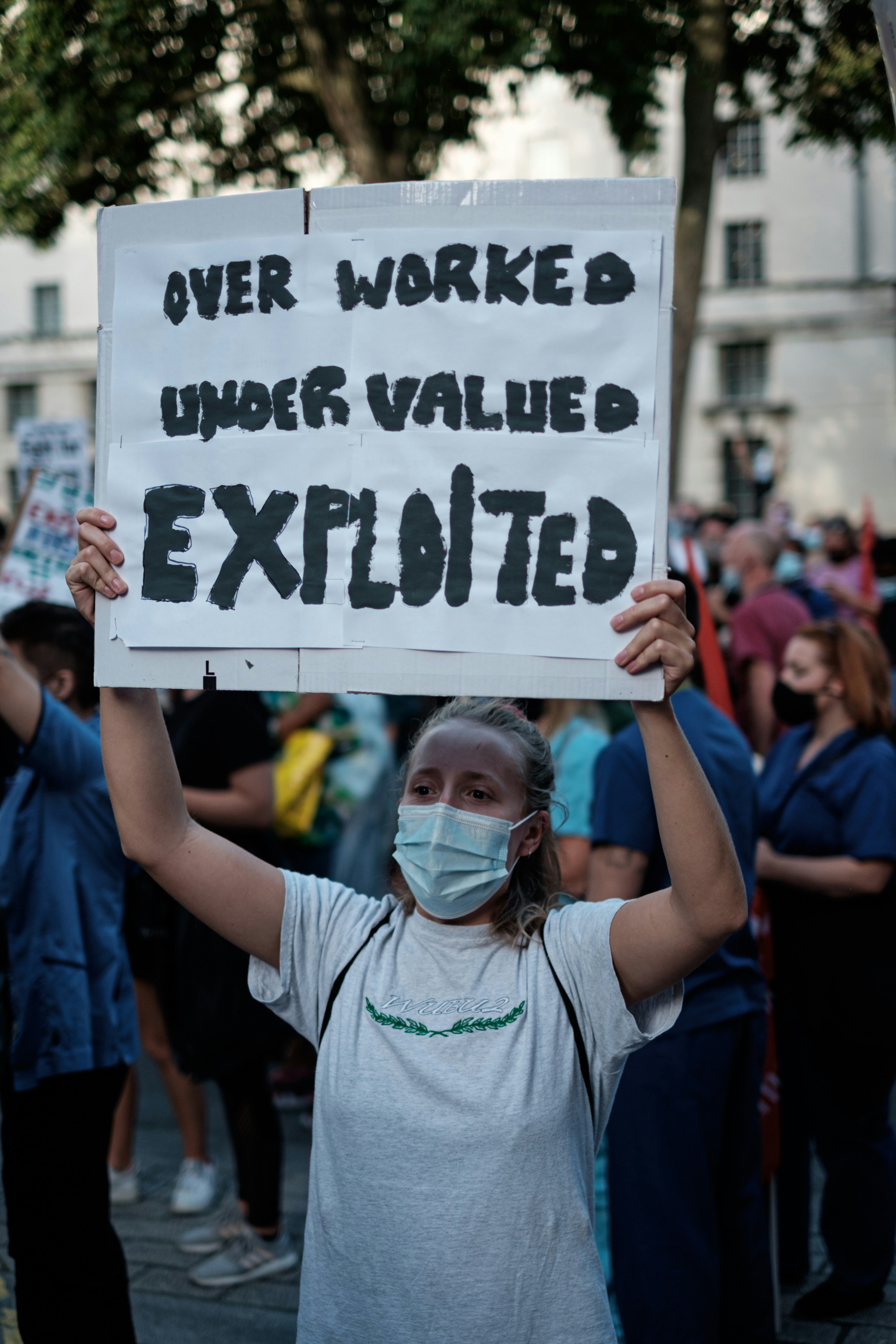 Eine Pflegekraft hält ein Schild in die Höhe auf dem steht: OVER WORKED UNDER VALUED EXPLOITED zu deutsch: ÜBERARBEITET UNTERBEZAHLT AUSGEBEUTET