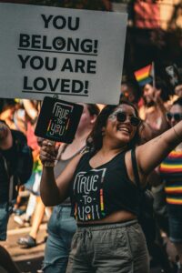 Das Foto zeigt eine Person, die bei einer bunten Demonstration mitläuft. Die Person hält ein großes Schild über ihren Kopf, auf dem steht: „YOU BELONG! YOU ARE LOVED!“ (deutsch: Du gehörst dazu! Du bist geliebt!). Sie hält ein weiteres kleineres Schild, auf dem „TRUE TO YOU“ steht. Die Person trägt ein schwarzes Top mit demselben Aufdruck. Im Hintergrund sind weitere Menschen zu sehen. Viele tragen bunte Kleidung, Regenbogenfarben und halten kleine inklusive Regenbogenfahnen. Die Stimmung wirkt offen, positiv und unterstützend, als stünde die Gruppe für Selbstakzeptanz und gegenseitige Wertschätzung ein.