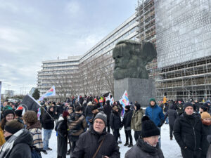 Das Bild zeigt eine Demonstration oder Kundgebung auf einem Platz mit winterlicher Kulisse – es liegt Schnee. Im Zentrum befindet sich das berühmte Karl-Marx-Monument, ein riesiger Kopf aus Bronze auf einem steinernen Sockel. Rund um das Denkmal stehen viele Menschen, die sich warm angezogen haben. Sie halten Fahnen und Banner in der Hand, auf denen verschiedene Symbole und Farben zu erkennen sind. Im Hintergrund sind hohe, modernistische Plattenbauten sowie ein großes, eingerüstetes Gebäude zu sehen, dessen Fassade mit riesigen Inschriften versehen ist.
