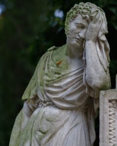 Das Bild zeigt eine alte Steinstatue, die eine Figur in klassisch drapierter Gewandung darstellt. Die Statue ist teilweise von grünem Moos oder Algen überzogen, was auf das Alter und die Witterungseinflüsse hindeutet. Die Figur steht an einem kunstvoll verzierten steinernen Sockel oder Grabmal. Die rechte Hand der Statue ist an den Kopf gelegt, was eine Geste der Trauer oder Besinnung ausdrücken könnte. Das Gesicht der Statue ist auf dem Bild unkenntlich gemacht. Im Hintergrund ist unscharfes, grünes Laub zu erkennen, was auf einen Standort im Freien, vermutlich auf einem Friedhof oder in einem Park, schließen lässt. Die Atmosphäre wirkt ruhig, nachdenklich und leicht melancholisch.
