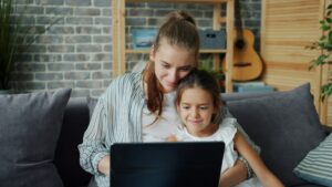 Das Bild zeigt eine erwachsene Person und ein Kind, die gemeinsam auf einem Sofa sitzen. Sie sind einander zugewandt und schauen zusammen auf einen Laptop, der vor ihnen auf dem Schoß der erwachsenen Person steht. Beide sind entspannt und kuscheln dicht beieinander. Im Hintergrund sieht man eine gemütliche Wohnzimmerszenerie mit grauen Kissen, einer Backsteinwand, Regalbrettern und einer Gitarre. Die Atmosphäre wirkt ruhig, familiär und gemütlich.