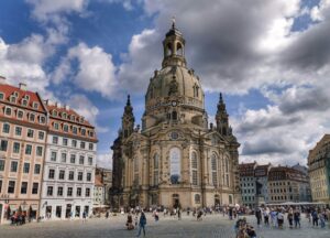 Das Bild zeigt einen belebten Platz in einer historischen Altstadt, im Zentrum steht die imposante Frauenkirche in Dresden. Die Kirche dominiert das Bild mit ihrer prächtigen barocken Architektur, der großen steinernen Kuppel und den zahlreichen verzierten Fassadendetails. Links und rechts der Kirche schließen sich farbenfrohe, mehrstöckige Altstadthäuser an, die in Pastelltönen gestrichen sind und harmonisch zum Stadtbild passen. Auf dem Platz vor der Kirche herrscht reges Treiben: Viele Menschen spazieren, stehen zusammen oder fotografieren die Sehenswürdigkeit. Am Himmel ziehen dramatisch wirkende Wolken vorbei, durch die stellenweise die Sonne scheint.