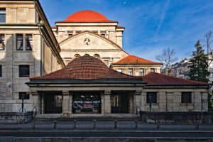 Das Bild zeigt die Synagoge in Frankfurt mit sandfarbenem Stein und roten Ziegeldächern. Der Baustil ist monumental und erinnert an klassische öffentliche Bauwerke. Im Zentrum des Bildes, am unteren Teil des Gebäudes, befindet sich ein Eingangsbereich mit einem kleinen Vorplatz. Über dem Eingang hängt ein großes Banner mit der Aufschrift „BRING THEM HOME! NOW“. Vor dem Banner, direkt am Boden, sind kleine Gestecke oder Blumen zu sehen.