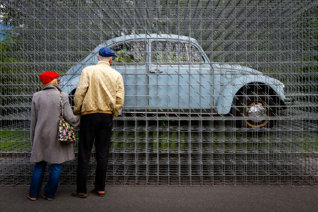 Das Bild zeigt zwei Personen, die von hinten zu sehen sind. Sie betrachten gemeinsam ein hellblaues, klassisches Auto (einen VW Käfer), das von einem großen Gitter aus Metallstäben vollständig umgeben ist. Das Auto scheint regelrecht im Gitter „gefangen“ zu sein. Die Szenerie spielt draußen.