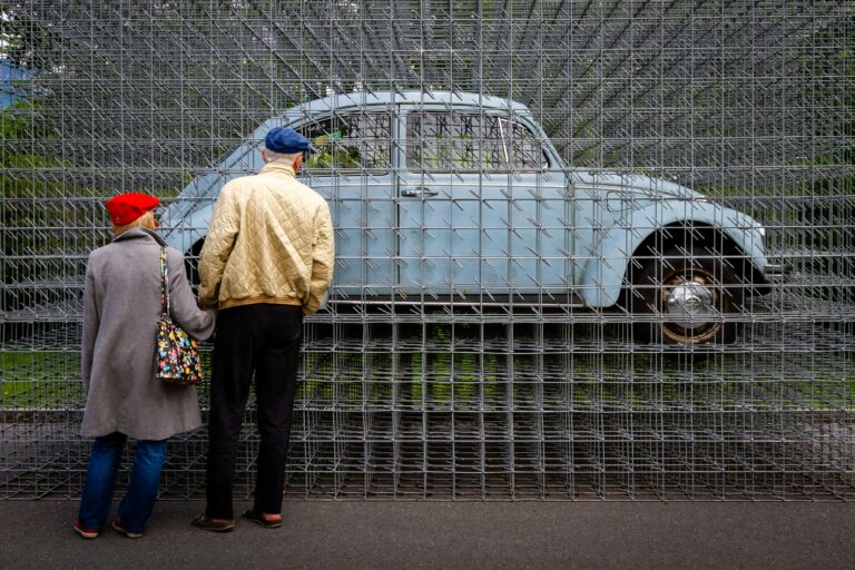 Das Bild zeigt zwei Personen, die von hinten zu sehen sind. Sie betrachten gemeinsam ein hellblaues, klassisches Auto (einen VW Käfer), das von einem großen Gitter aus Metallstäben vollständig umgeben ist. Das Auto scheint regelrecht im Gitter „gefangen“ zu sein. Die Szenerie spielt draußen.