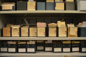 Ein Regal mit mehreren Reihen älterer Archivschachteln und Kartons, beschriftet mit Buchstaben und Jahreszahlen, jeweils mit farbigen Markierungen versehen. Dazwischen liegen lose Papiere und Aktenblätter. Offenbar handelt es sich um historisch geordnete Dokumentenbestände