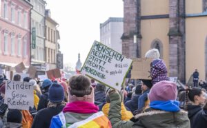 Das Bild zeigt eine Demonstration auf einer Straße in einer Stadt. Viele Menschen, fast alle warm angezogen mit Mützen und Winterjacken, sind von hinten zu sehen. Einige Teilnehmende halten Schilder in die Höhe. Auf einem Schild steht groß zu lesen: „MENSCHEN RECHTE STATT RECHTE MENSCHEN.“, daneben Hashtags wie #fck12 und #fckafd. Ein weiteres Schild, das teilweise sichtbar ist, trägt die Aufschrift „Kinder gegen Rechts“.