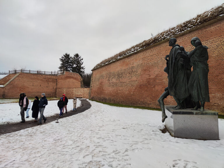 Eine Gruppe von Teilnehmenden bewegt sich an einem winterlichen Tag entlang der historischen Mauern der Kleinen Festung in Theresienstadt. Eine Skulptur im Vordergrund erinnert an die Geschichte des Ortes und verleiht der Szene eine stille, nachdenkliche Atmosphäre