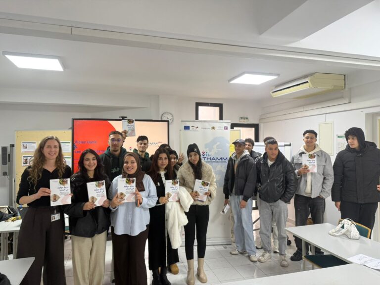 Das Bild zeigt eine Gruppe von etwa zwölf jungen Menschen in einem Seminarraum. Sie stehen nebeneinander, teilweise auch leicht hintereinander, und halten jeweils die Broschüre in der Hand. Die Menschen lächeln und wirken fröhlich. Im Hintergrund sind mehrere Informationsplakate sichtbar, unter anderem mit dem Logo „THAMM“.