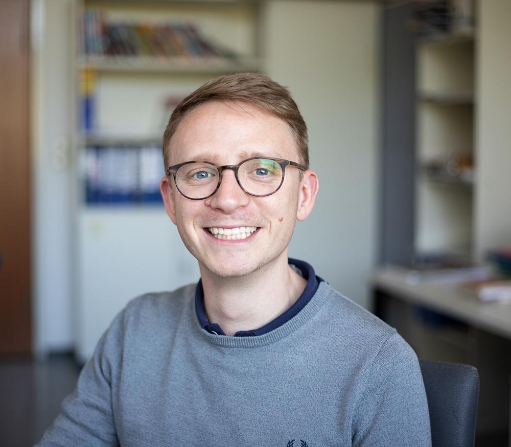 Eine Person sitzt in einem Büro; im Hintergrund sind Regale und Bücher zu sehen. Die Person trägt eine Brille und lächelt freundlich.