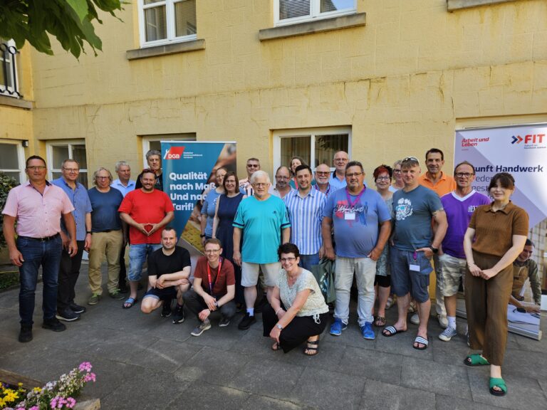 Eine Gruppe von etwa zwanzig Menschen steht und sitzt für ein Gruppenfoto im Hof vor einem gelben Gebäude. Im Hintergrund sind zwei Roll-Ups mit Logos und Texten zum Thema Handwerk und Tarif zu sehen. Die Stimmung wirkt freundlich und sommerlich.