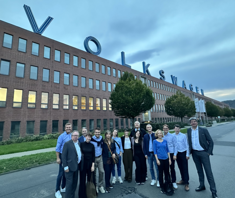 Eine Gruppe Menschen steht vor einem Gebäude der Firma Volkswagen. Sie lächeln freundlich in die Kamera.
